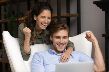 Happy young bonding family couple looking at laptop screen, celebrating getting email with good news. Emotional laughing spouses making yes gesture, reading amazing online lottery win notification.