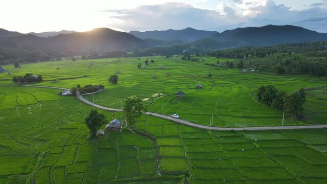 Aerial view slide camera parallel to the path into the farmer village amid green rice terrace fields during sunset over the mountain.