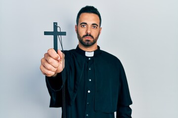 Handsome hispanic priest man with beard holding catholic cross thinking attitude and sober...