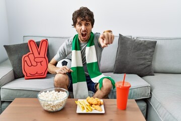 Young hispanic man football hooligan cheering game at home pointing displeased and frustrated to the camera, angry and furious with you