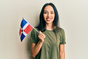 Young hispanic girl holding dominican republic flag looking positive and happy standing and smiling with a confident smile showing teeth