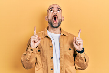 Young bald man wearing casual clothes amazed and surprised looking up and pointing with fingers and...