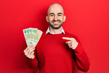 Young bald man holding argentine pesos banknotes smiling happy pointing with hand and finger