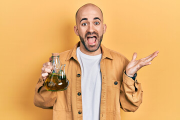 Young bald man holding olive oil can celebrating achievement with happy smile and winner expression...