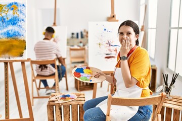 Young artist woman painting on canvas at art studio laughing and embarrassed giggle covering mouth...