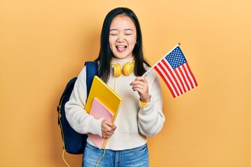 Young chinese girl exchange student holding america flag sticking tongue out happy with funny expression.