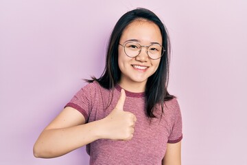 Fototapeta premium Young chinese girl wearing casual clothes and glasses doing happy thumbs up gesture with hand. approving expression looking at the camera showing success.
