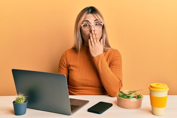 Young caucasian woman working at the office wearing glasses covering mouth with hand, shocked and afraid for mistake. surprised expression