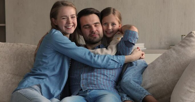 Beautiful young couple with daughter sit on sofa cuddling smiling look at camera. Loving woman her daughter embracing beloved husband dad express love, congratulate Happy Fathers Day. Bonding concept
