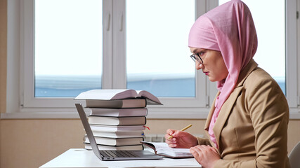 Muslim woman writes with a pen in a notebook looking into a laptop.