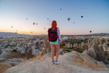 Girl with a backpack posing against the backdrop of a valley with mountains and balloons at sunrise. Back view. Entertainment, tourism an vacation. Travel tour. Goreme, Cappadocia, Turkey.