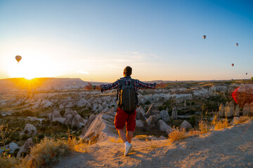 Back view. Man with arms spread apart enjoy the sunrise. Tourist attraction balloon flight on background. Entertainment, tourism an vacation. Travel tour. Goreme, Cappadocia, Turkey.