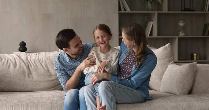 Lovely 8s little daughter sitting on loving parents laps spend time together at modern home, resting on cozy sofa in living room. Happy parenthood, homeowners family moments, adoption, custody concept