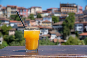 A glass of freshly squeezed orange juice stands on a wooden table top against the background of the city on a sunny summer day.