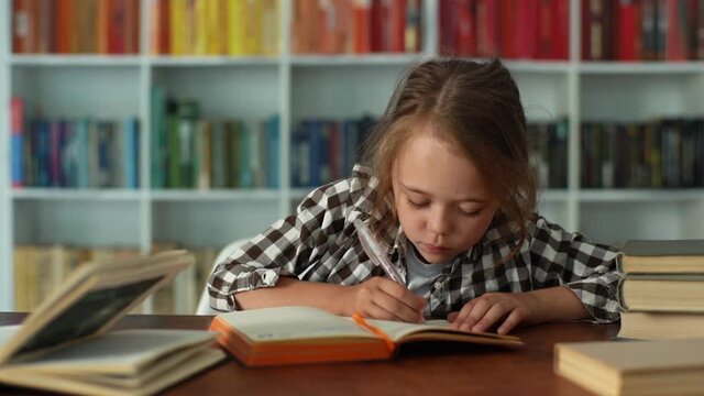 Close-up front view of happy pretty elementary child school girl finishing homework and closing workbook sitting at desk. Adorable smiling primary pupil schoolgirl completing exercise in copybook.