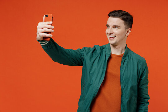 Smiling beautiful fun happy young brunet man 20s wears red t-shirt green jacket doing selfie shot on mobile cell phone post photo on social network isolated on plain orange background studio portrait.