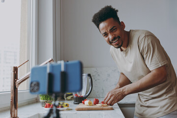 Young confused african american man in casual clothes prepare sweet breakfast cut fruit watch series on mobile cell phone cooking food in indoor kitchen at home alone Healthy diet lifestyle concept