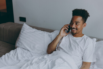 Young happy satisfied african american man in nightwear speak mobile cell phone lying in bed rest relax spend time in bedroom lounge home in own room house wake up dream be lost in reverie good day.