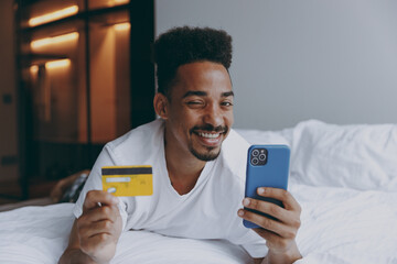 Full size happy young african american man in nightwear hold mobile cell phone credit bank card shopping online order delivery lying in bed rest spend time in bedroom home in own room house wake up