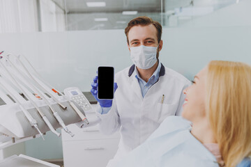Young friendly man dentist show mobile cell phone with blank screen area near patient woman with...