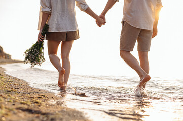 Cropped close up back view young couple two family man woman in white clothes hold flowers bouquet hand walk in water waves at sunrise over sea beach ocean outside seaside in summer day sunset evening