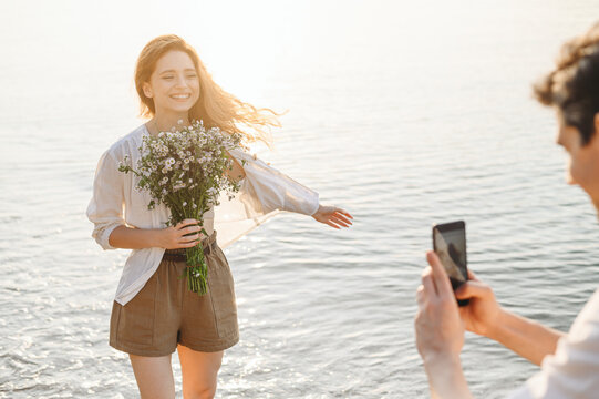 Sunlit young couple family man woman in white clothes hold flowers bouquet take photo on mobile cell phone rest together at sunrise over sea beach ocean outdoor seaside in summer day sunset evening