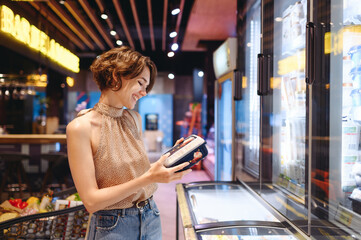 Young woman in casual clothes shopping at supermaket grocery store buy choose products stand near refrigerator hold container read ingredients shelf life inside hypermarket. Purchasing food concept.