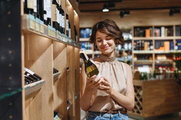 Young satisfied woman 20s in casual clothes shopping at supermaket grocery store buy choosing white wine alcohol hold bottle prop up chin inside hypermarket. People purchasing gastronomy food concept.