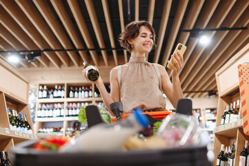 Young satisfied fun woman in casual clothes shopping at supermaket store with grocery cart hold white wine alcohol hold bottle talk speak mobile phone inside hypermarket Purchasing gastronomy concept