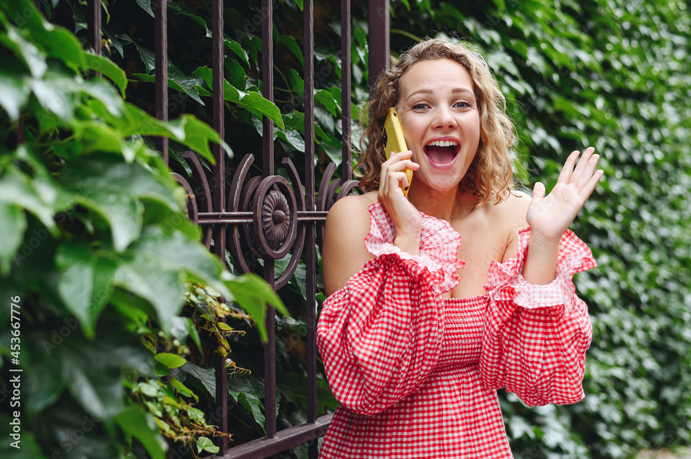 Wall mural Surprised young freelancer caucasian smiling happy fun amazed woman 20s wearing pink dress talk on mobile cell phone spread hand walk in city standing outdoor near forged gate on green ivy background