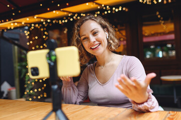 Young smiling fun happy woman 20s wearing casual clothes sit alone at table in cafe shop listen music in earphones mobile cell phone talk by video call do selfie shot relaxing in restaurant indoors.