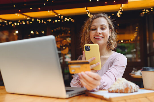Young woman in casual clothes sit at table in cafe shop eat breakfast using laptop pc computer take photo of credit bank card by mobile phone shopping online order delivery relax in restaurant indoor