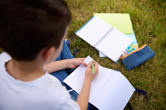 Overhead View Of School Boy Writing On Empty Blank Sheets Of A Workbook. Rear View Of An Elementary Aged Child Doing Homework School Task At The Park After School . School Supplies Lying On Grass