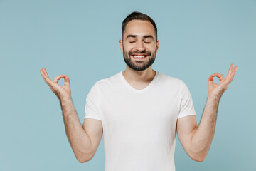 Young spiritual man in blank print design white t-shirt hold spread hands in yoga om aum gesture relax meditate try to calm down isolated on plain pastel light blue color background studio portrait