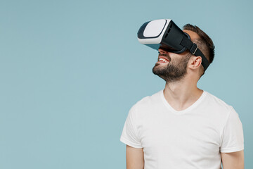 Young happy excited amazed man 20s wear casual basic white t-shirt play watching in vr headset pc gadget isolated on plain pastel light blue color background studio portrait. People lifestyle concept.