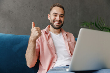 Young satisfied happy smiling smart man 20s wear casual clothes hold use work on laptop pc computer show thumb up like approve gesture sitting on blue sofa at home flat indoors rest relax on weekends