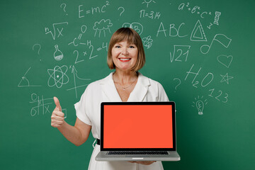 Teacher mature elderly woman 55 wear white shirt hold use laptop pc computer with blank screen workspace area show thumb up like gesture isolated on green wall chalk blackboard background studio