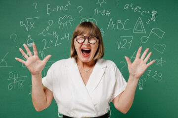 Angry teacher mature elderly lady woman 55 wear shirt glasses spreading hands scream eyes close isolated on green wall chalk blackboard background studio. Education in high school September 1 concept