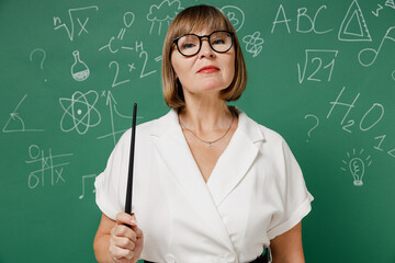 Serious clever teacher mature elderly lady woman 55 wear shirt glasses holding pointer in hand isolated on green wall chalk blackboard background studio. Education in high school September 1 concept.