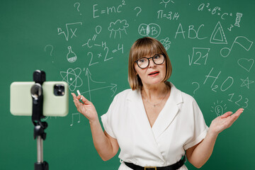 Clever teacher mature elderly lady woman 55 wear shirt glasses work with mobile cell phone on tripod on lockdown quarantine spreading hands isolated on green wall chalk blackboard background studio.