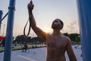 Shirtless Latinx man adjusting a resistance band, preparing for an outdoor workout at sunset