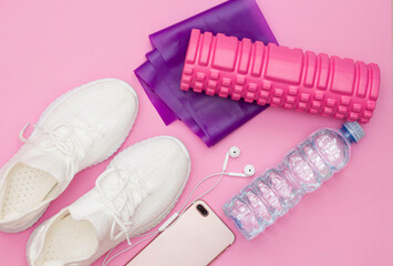 A fitness concept with a water bottle, sneakers, a phone with headphones, a purple fitness elastic band, a pink roller on a pink background. Copy space.