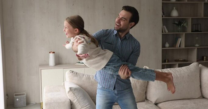 Cheerful father swings on hands cute 9s daughter, girl spread arms like plane wings imagines flying in air. Family dreaming about travel and holidays. Leisure activities with children at home concept