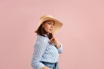 Obraz na płótnie Canvas Sexy woman with dark hair makes shooting gesture at camera, wants you, poses in straw hat, flirts, feels sexy, wears striped shirt, jeans with belt, on pink background. Summer emotions concept.