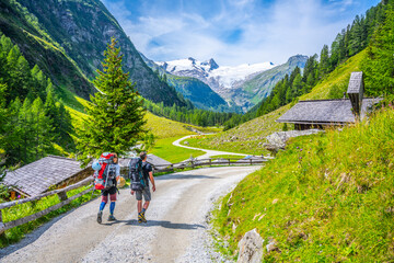 Two backpackers hike on the country road towards high mountains. Grossvenediger in Hohe Tauern...