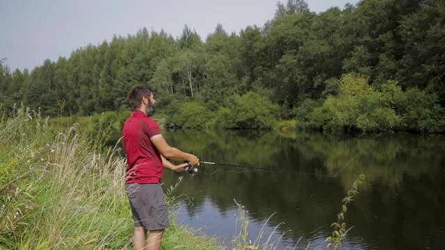 A man with a beard is fishing on the river, casting a line. A fisherman with a fishing rod is fishing on the river bank.