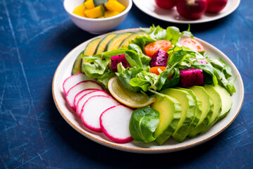Healthy vegan salad fruits and vegetables dish with avocado, dragon fruit, radish, pumpkin, tomato, corn salad, lettuce, mizuna and lemon