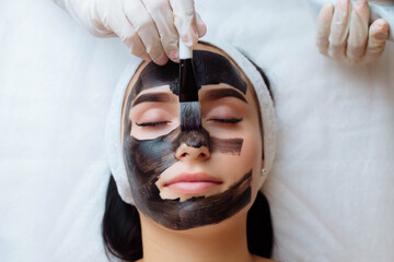 cosmetologist applying black mask on pretty woman face wearing black gloves, gorgeous woman in spa having facial procedures