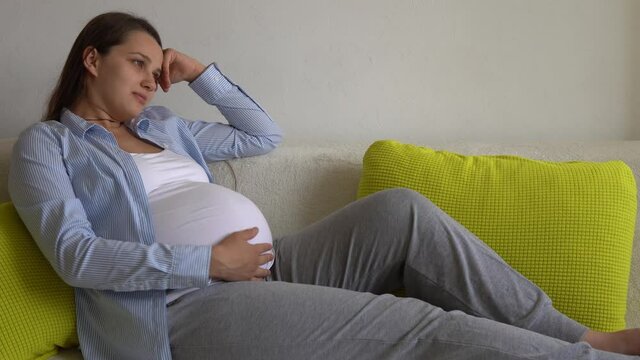 Cute Pregnant Woman strokes large belly touching tummy in sunny day. Young Ledy In Pregnancy Have Fun Spend Time On yellow Sofa At Home. Motherhood, Parenthood, Medicine Health and Care prenatal