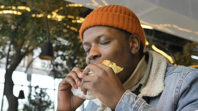 Black guy in jacket and hat tastes delicious burger sitting on fast food restaurant terrace against blurred trees close view